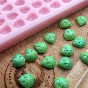 May include: A pink silicone mould with 36 cavities shaped like alien heads. The mould is next to a wooden surface with 15 green alien head shaped sweets.