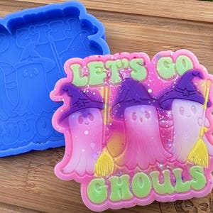 May include: A pink and green resin mold with three ghosts wearing witch hats and holding brooms. The mold says "Let's Go Ghouls".