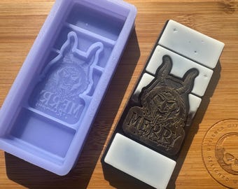 Könnte beinhalten: Eine lilafarbene Silikonform mit einem Design einer Kreatur mit Hörnern und dem Wort "Merry" in gotischer Schrift. Die Form liegt neben einer schwarz-weißen Seifenstange mit dem gleichen Design.