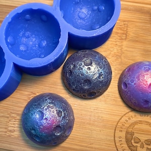 Pode incluir: Um molde de silicone azul com três cavidades esféricas está ao lado de três esferas decorativas. As esferas são pintadas com cores metálicas e têm uma superfície craterada. Uma esfera é roxa e rosa, outra azul e prateada, e a terceira é multicolorida.