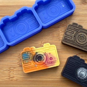 May include: A silicone mould for making camera-shaped soaps or resin crafts. Three finished cameras are shown in the image: one with a brown wood-like finish, one with a black finish, and one with a yellow, orange, and red finish with colourful speckles.