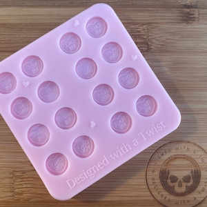 Puede incluir: Molde de silicona rosa con 16 cavidades en forma de corazón. Cada cavidad tiene un mensaje diferente, como "Kiss Me", "Love Bug" y "Be Mine". El molde está diseñado con un toque de originalidad y tiene el texto "Designed with a Twist" en la parte inferior.