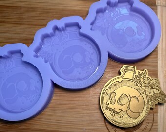 Pode incluir: Três moldes de silicone roxo para fundição de resina, cada um com um design redondo de uma garrafa de vidro com uma caveira dentro e detalhes florais. Os moldes são projetados para criar um pingente ou um amuleto. O texto "DESIGNED BY LITTLE" é visível em um dos moldes. Também é mostrado um pingente de metal dourado com o mesmo design.