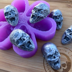 Puede incluir: Moldes de resina plateada con forma de calaveras de zombi. Las calaveras están dispuestas sobre un molde morado con forma de flor y una superficie de madera. Las calaveras tienen la boca abierta y rasgos faciales detallados. El tema es el terror y Halloween.