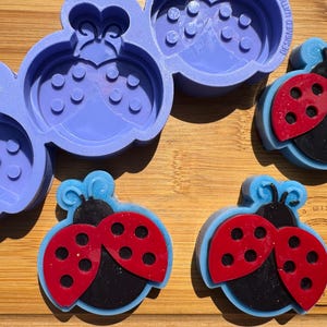 May include: A close-up of a wooden surface with a purple ladybug mold and several finished ladybug-shaped objects. The ladybugs are red and black with blue accents. The mold is made of flexible silicone.