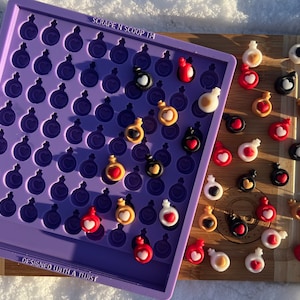 May include: A purple silicone mould with potion bottle-shaped cavities, filled with colourful resin charms. The charms are red, black, white, and gold, each with a heart design. The mould is on a wooden chopping board. The text "SCRAPE N SCOOP" is visible.