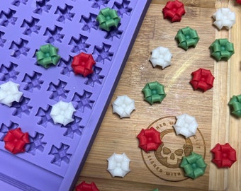 Pode incluir: Uma forma de silicone roxa com uma grade de 36 cavidades em forma de estrela. A forma está cheia de pequenos doces em forma de laço, de cores vivas. Os doces são vermelhos, verdes e brancos. Os doces estão dispostos sobre uma superfície de madeira.