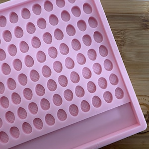 May include: A light pink silicone mold with numerous oval-shaped cavities, each featuring a textured pattern. The mold is rectangular with a smooth, flat base and is set against a wooden cutting board.
