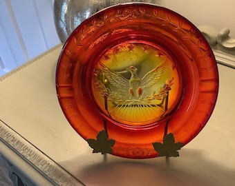 May include: A red and orange glass plate with an embossed eagle design. The plate is on a decorative stand with leaf-shaped accents.