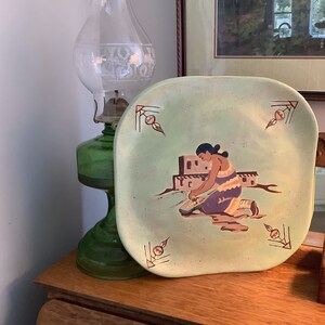 May include: A green ceramic plate with a painted design of a woman grinding corn in front of a pueblo-style building. The plate has a light green background with brown, orange, and purple accents.