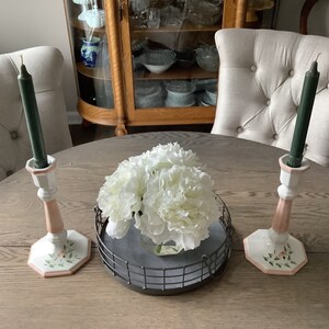 May include: A round wooden table with two white chairs and a centerpiece of white flowers in a glass vase on a black metal tray. Two white ceramic candle holders with pink accents and green candles are on the table.