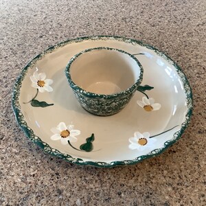 May include: A white ceramic serving dish with a green rim and a smaller bowl in the center. The dish is decorated with white daisies with yellow centers.