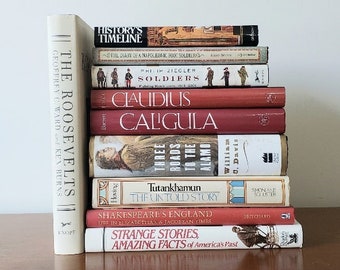 May include: A stack of hardcover books with various titles, including "The Roosevelts," "History's Timeline," and "Strange Stories." The books are in shades of red, white, and brown, arranged on a wooden surface.