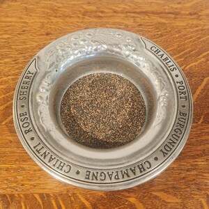 May include: A silver-colored ashtray with a circular design. The rim is embossed with grape clusters and the names of wines: Sherry, Chianti, Rose, Chablis, Port, Burgundy, and Champagne. The center has a textured, dark brown surface.
