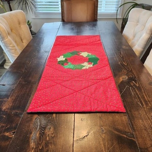 May include: Red quilted table runner with a green and gold wreath design. The runner is on a dark wood table.