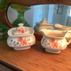May include: A white sugar bowl and creamer set with a floral design featuring orange and green flowers. The set is on a wooden surface.