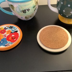 Pu&ograve; includere: Due sottobicchieri, uno con un design floreale e un bordo arancione, l'altro con un top in sughero e un bordo bianco.