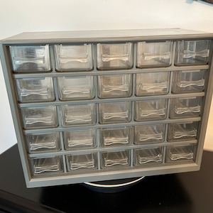 May include: A gray plastic drawer organizer with 24 clear plastic drawers. The organizer is sitting on a black surface.