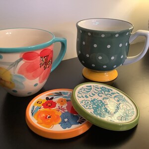 Pu&ograve; includere: Due tazze in ceramica con motivi floreali e a pois, e due sottobicchieri abbinati con motivi floreali. Le tazze sono entrambe bianche con motivi colorati. I sottobicchieri sono rotondi e hanno un bordo verde.