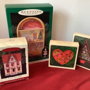 May include: A set of five Hallmark Keepsake Christmas ornaments. The ornaments are packaged in their original boxes. The ornaments include a Victorian Painted Cottage, a heart-shaped ornament with red and gold embroidery, a Christmas tree ornament with black and gold embroidery, a miniature doll in a pink dress, and a miniature dollhouse with a Christmas tree inside.