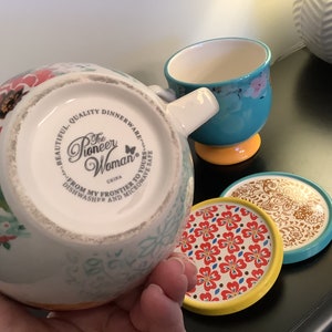 May include: A white ceramic mug with a floral design and a turquoise rim. The mug has a handle and the words "The Pioneer Woman" printed on the bottom. The mug is sitting on a coaster with a floral design.