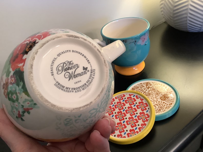 May include: A white ceramic mug with a floral design and a turquoise rim. The mug has a handle and the words "The Pioneer Woman" printed on the bottom. The mug is sitting on a coaster with a floral design.