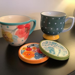Pu&ograve; includere: Due tazze in ceramica con disegni floreali e a pois, e due sottobicchieri abbinati con motivi floreali. Le tazze sono sopra i sottobicchieri.