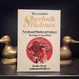 May include: A vintage book titled "The Complete Sherlock Holmes" by Sir Arthur Conan Doyle. The cover features a red and white illustration of a hat, magnifying glass, and candle. The book is an authorized edition by Berkley Books.