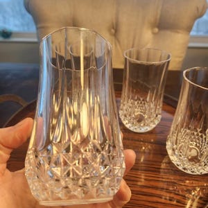 May include: A set of four clear crystal drinking glasses. The glasses have a diamond-cut pattern on the lower half and a smooth, slightly flared rim. The glasses are displayed on a wooden tray.