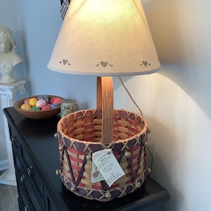 May include: A woven basket lamp with a wooden handle and a white lampshade with brown hearts. The basket is decorated with brass bells and has a red, brown, and white woven pattern. The lamp is sitting on a black dresser.
