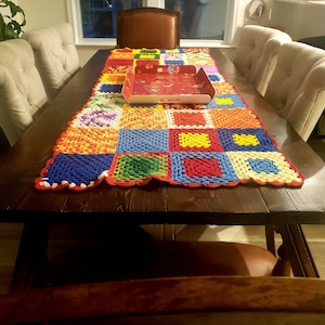 May include: A colorful crocheted table runner with a patchwork design in shades of red, orange, yellow, green, blue, and purple. The runner is laid out on a dark brown wooden table with six chairs.