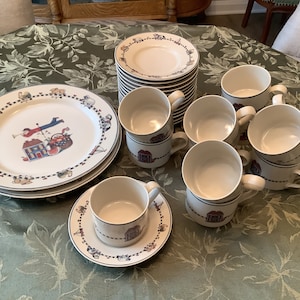 May include: A complete cream-colored dinnerware set with a whimsical design. The set includes plates, saucers, and mugs, each adorned with a blue border and a repeating pattern of houses and figures. The plates measure approximately 10 inches in diameter.