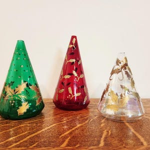 May include: Three cone-shaped glass ornaments. One is green with gold leaf designs, the second is red with gold leaf designs, and the third is clear with gold leaf designs. They are displayed on a wooden surface.