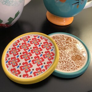 May include: Two round ceramic coasters with floral patterns. One coaster has a yellow rim and a red, white, and blue floral design. The other coaster has a teal rim and a brown floral design.