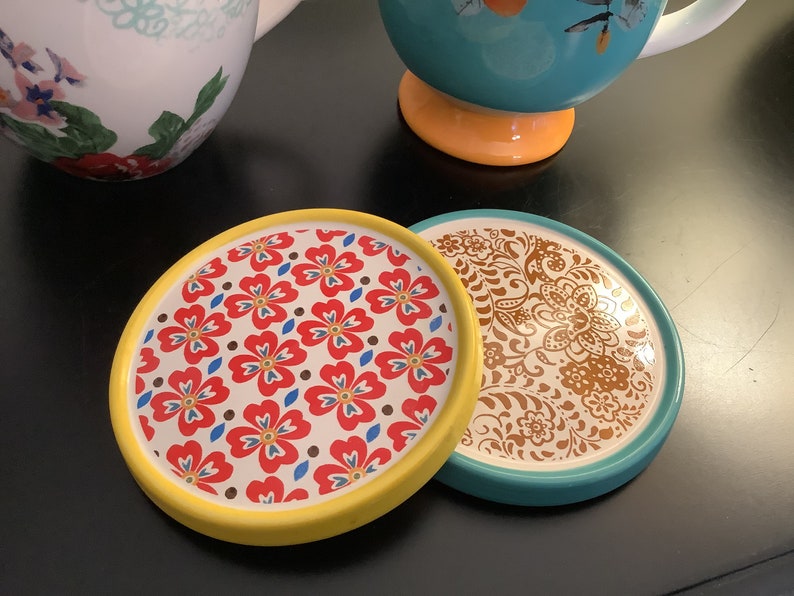 May include: Two round ceramic coasters with floral patterns. One coaster has a yellow rim and a red, white, and blue floral design. The other coaster has a teal rim and a brown floral design.