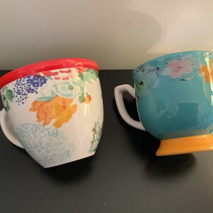 May include: Two ceramic mugs with floral patterns. One mug has a red rim and a white background with colorful flowers. The other mug has a turquoise body with a yellow base and a floral pattern in white, pink, and yellow.