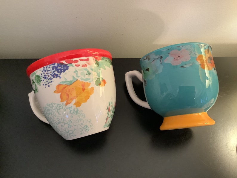 May include: Two ceramic mugs with floral patterns. One mug has a red rim and a white background with colorful flowers. The other mug has a turquoise body with a yellow base and a floral pattern in white, pink, and yellow.