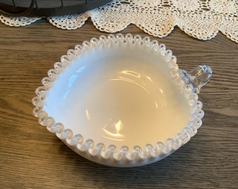 May include: A white milk glass bowl with a scalloped edge and a clear glass handle. The bowl has a shiny finish and is perfect for serving snacks or desserts.