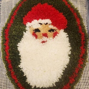 May include: Handmade Christmas rug featuring Santa Claus. The rug has a white beard, a red hat, and a tan face. The Santa Claus image is surrounded by a green and red border. The rug is made of yarn and is on a white mesh background.