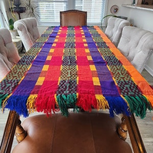 May include: A colorful woven table runner with fringe edges in shades of red, orange, yellow, green, purple, and blue. The runner is laid out on a wooden table with chairs around it.
