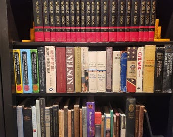 May include: A black bookshelf filled with various books. The top shelf features a set of red and black "World Book" encyclopedias. The middle shelf displays historical and biographical books, including titles like "Nelson" and "The Civil War".