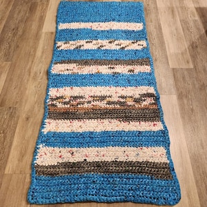 May include: A rectangular, handmade rug with a blue border and horizontal stripes. The stripes alternate between blue, white, gray, and a speckled mix of colors. The rug is made of a thick, textured material.