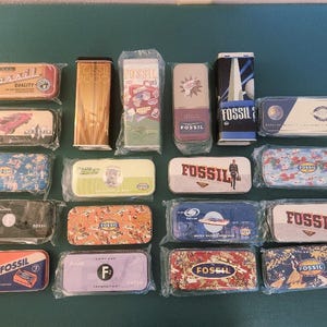 May include: A collection of Fossil brand metal tins in various shapes and designs. Some tins feature text like "Fossil" and "Start your next weekend with refreshing Fossil." The tins are in clear plastic packaging.