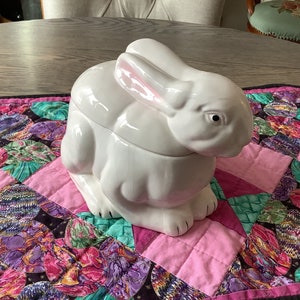 May include: White ceramic bunny shaped cookie jar with pink ears. The jar is sitting on a colorful patchwork quilt.