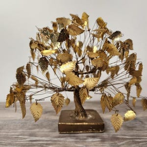 May include: A decorative tree sculpture featuring a twisted metal trunk and branches adorned with numerous small, gold-colored metallic leaves. The tree is mounted on a square, solid base, creating a structured, upright botanical form.