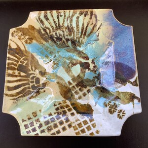 May include: A square ceramic dish with a unique design. The dish has a white background with brown, blue, and green patterns. The patterns are abstract and resemble footprints, leaves, and geometric shapes.