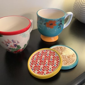 May include: Three ceramic mugs with floral patterns and three matching coasters. The mugs are teal, white with red trim, and white with pink trim. The coasters are yellow, white with red flowers, and brown with gold accents.