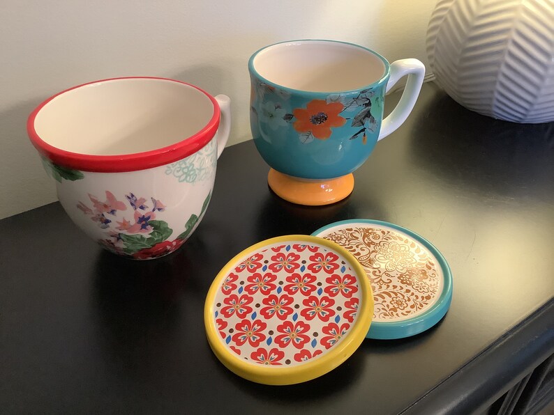 May include: Three ceramic mugs with floral patterns and three matching coasters. The mugs are teal, white with red trim, and white with pink trim. The coasters are yellow, white with red flowers, and brown with gold accents.