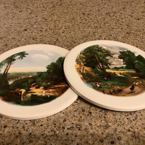 May include: Two round ceramic coasters with a white background and a painted landscape scene. The coasters feature a green, brown, and blue color scheme.
