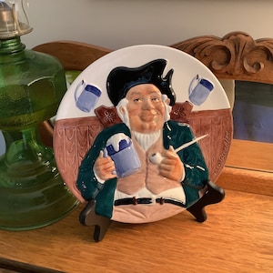 May include: A ceramic plate featuring a cartoon man with a pipe and a mug of beer. The plate is decorated with a wooden panel background and the text "Ciao Bella".
